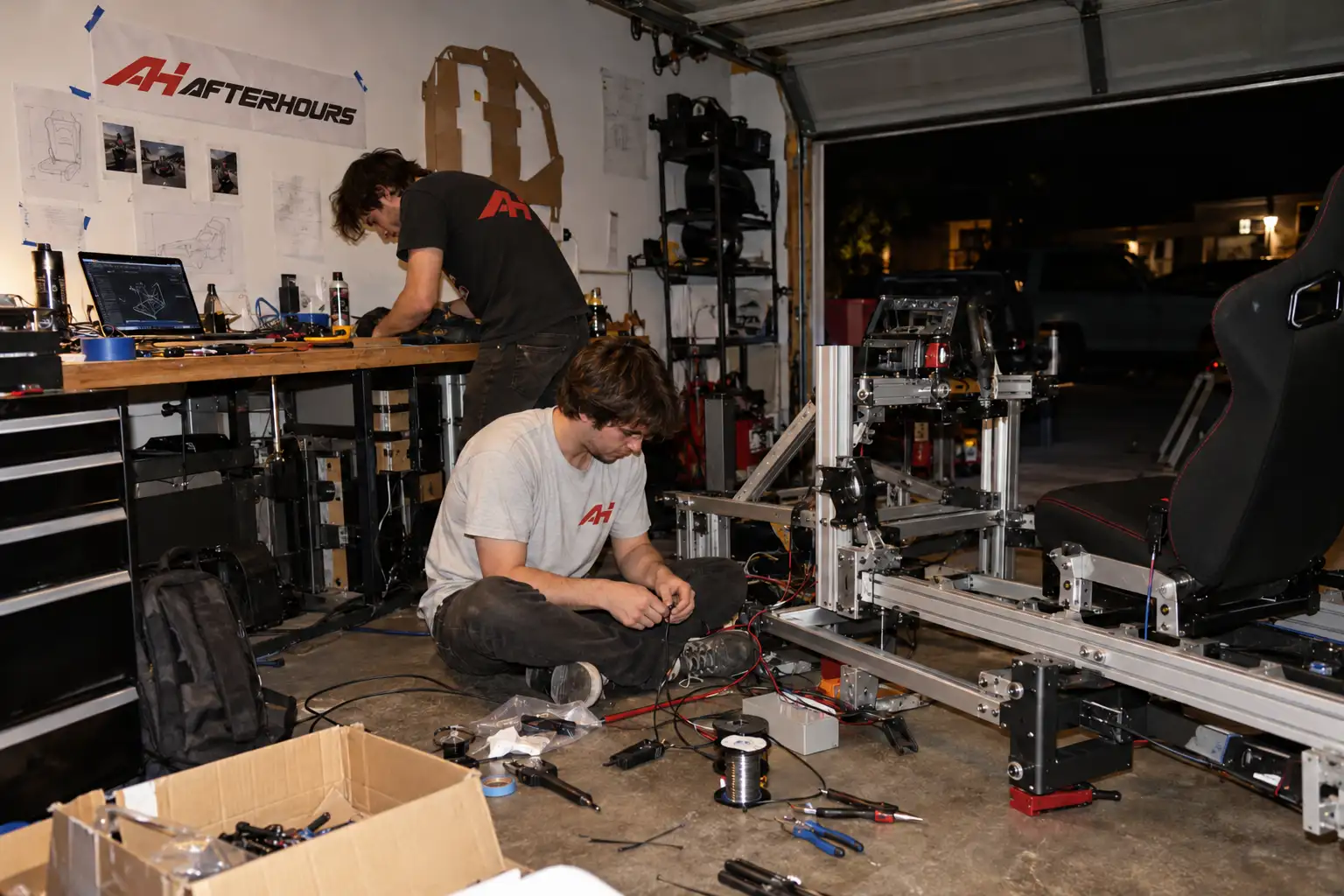 A garage workshop at night: two of the team build the first bare aluminium cockpit frame on the floor, sketches and the AH AFTERHOURS banner on the wall behind them