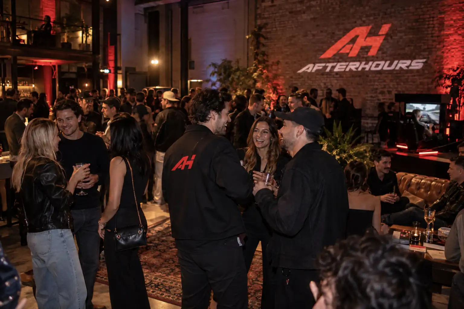 Launch night in a brick warehouse: a crowd with drinks, the AH monogram and the AFTERHOURS name projected on the wall, a simulator lit red on a low stage