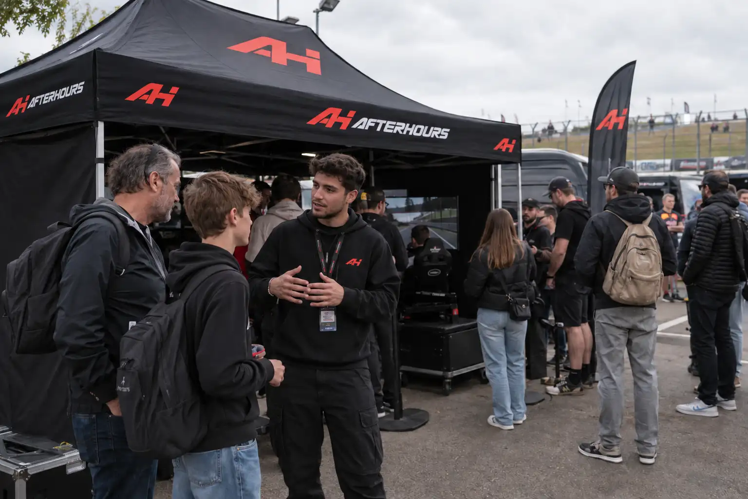 Under the black AFTERHOURS canopy in a circuit paddock, a member of the team in an AH hoodie talks with a father and son; a simulator runs inside the tent and visitors queue behind