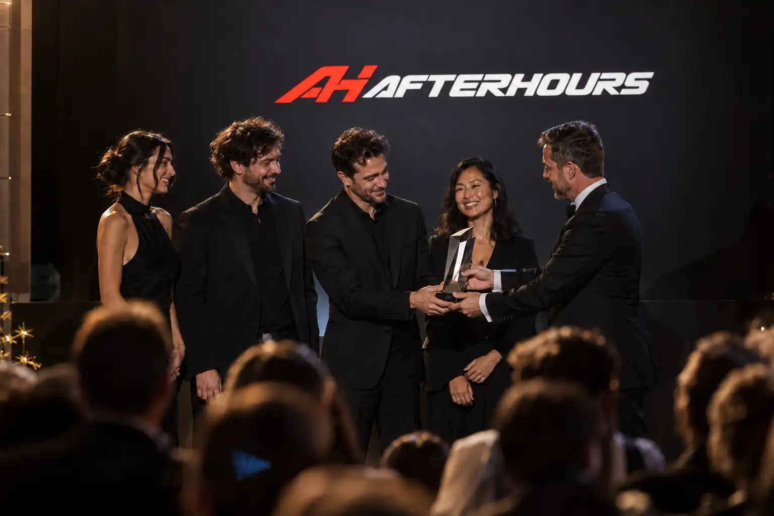 On stage in front of the AFTERHOURS name, four members of the team in black accept a crystal trophy from a presenter in a dinner jacket