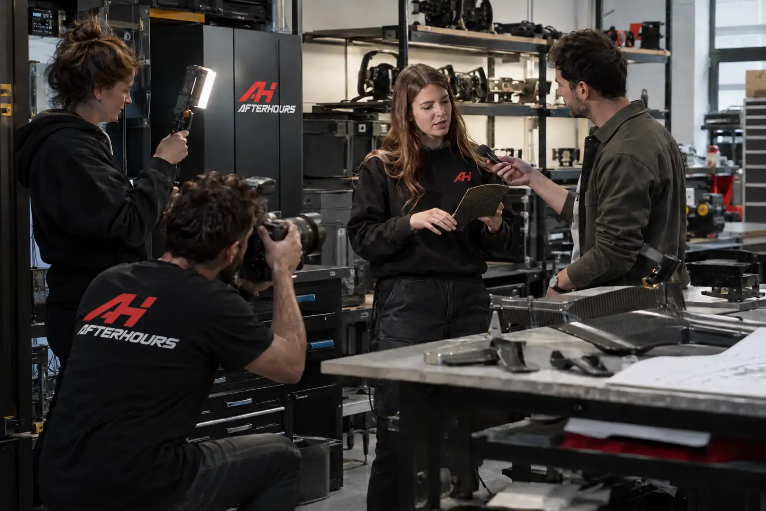 In the AFTERHOURS workshop, an engineer holds up a carbon panel while she is interviewed and photographed; wheels and pedal sets on the shelves behind her